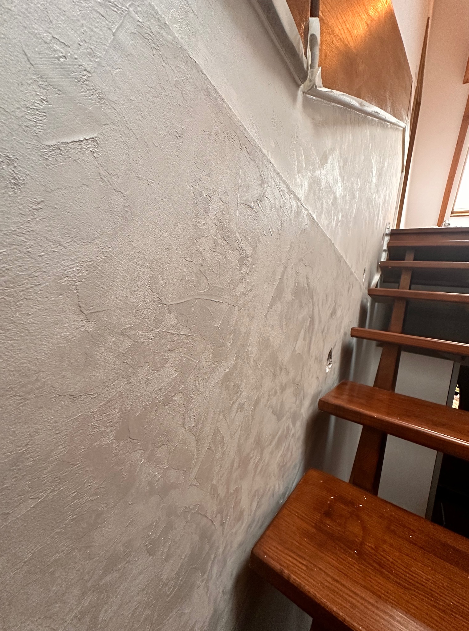 Dekoracyjna ściana z efektem stiuku w odcieniach szarości, sąsiadująca z drewnianymi schodami. Widoczne zabezpieczenie folią malarską.