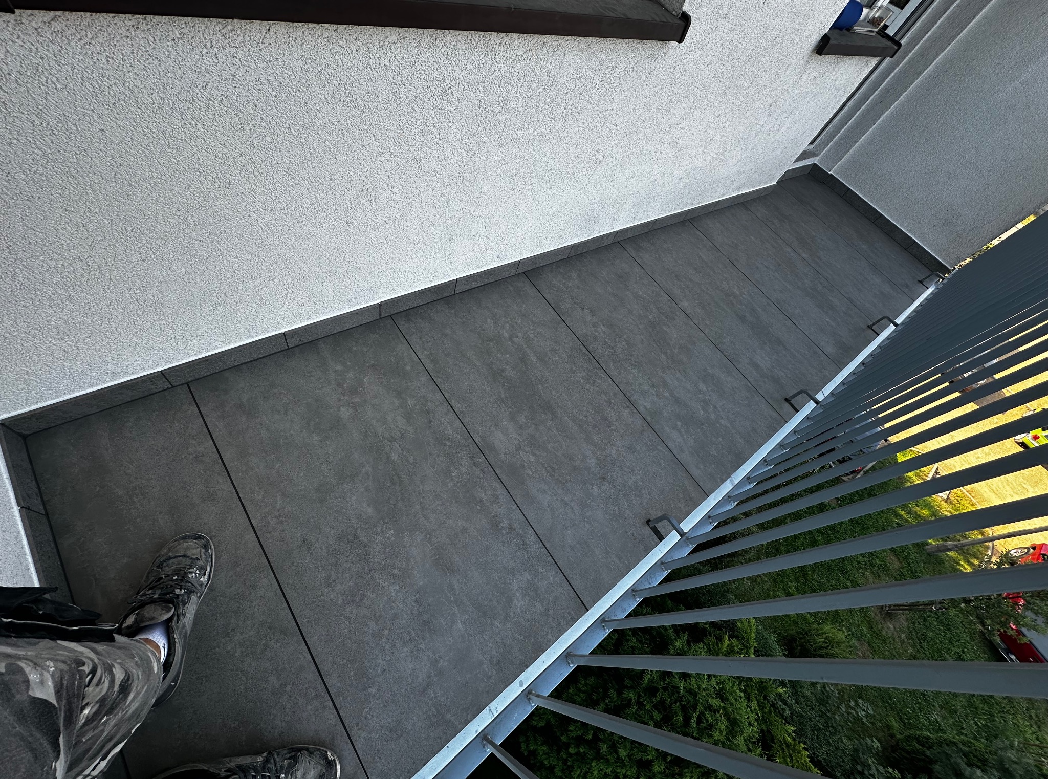 Szare płytki na balkonie ułożone równolegle do ściany, widok z góry, fragment balustrady i butów osoby wykonującej pracę. Widoczne połączenia między płytkami.