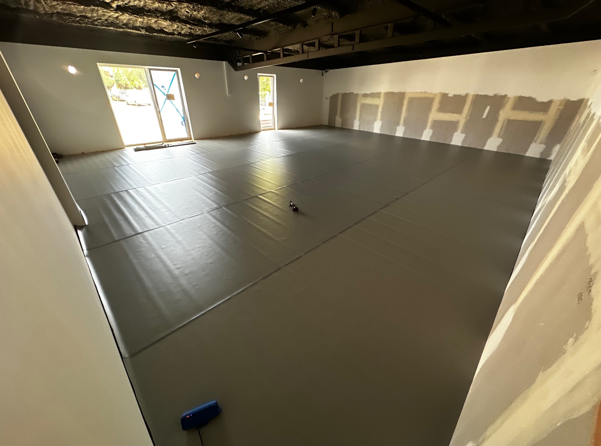 Sala z szarymi matami podłogowymi, przygotowana do ćwiczeń. Ściany częściowo wykończone, sufit w ciemnym kolorze. Widoczne okna z jasnym światłem dziennym.