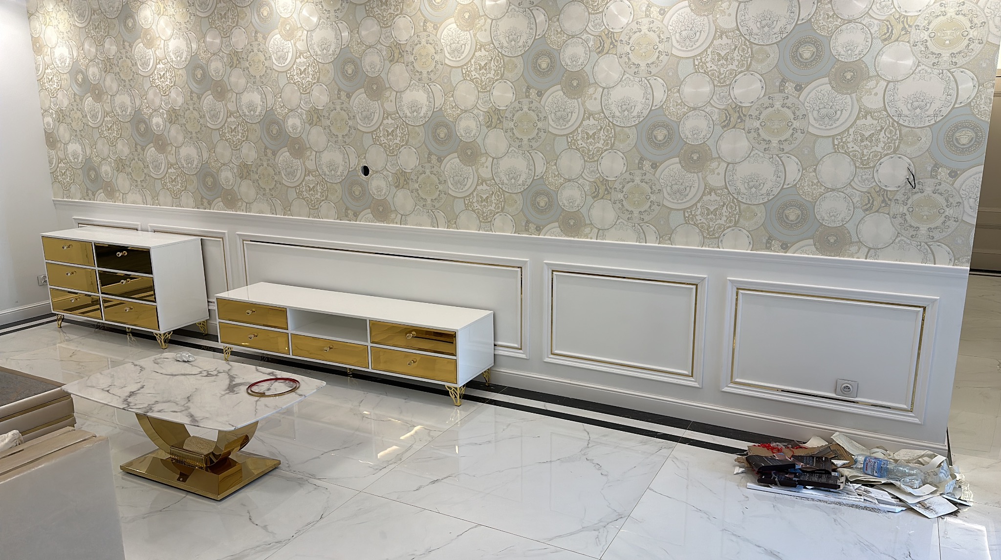 Eleganckie wnętrze: białe meble z złotymi akcentami na tle wzorzystej tapety. Marmurowa podłoga i stolik kawowy dopełniają luksusowy charakter pomieszczenia.