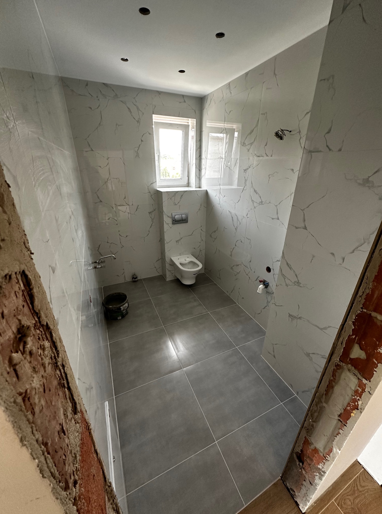 Wykończona łazienka z szarymi płytkami podłogowymi i białymi płytkami ściennymi imitującymi marmur, z oknem i częściowo wykończonym wejściem.