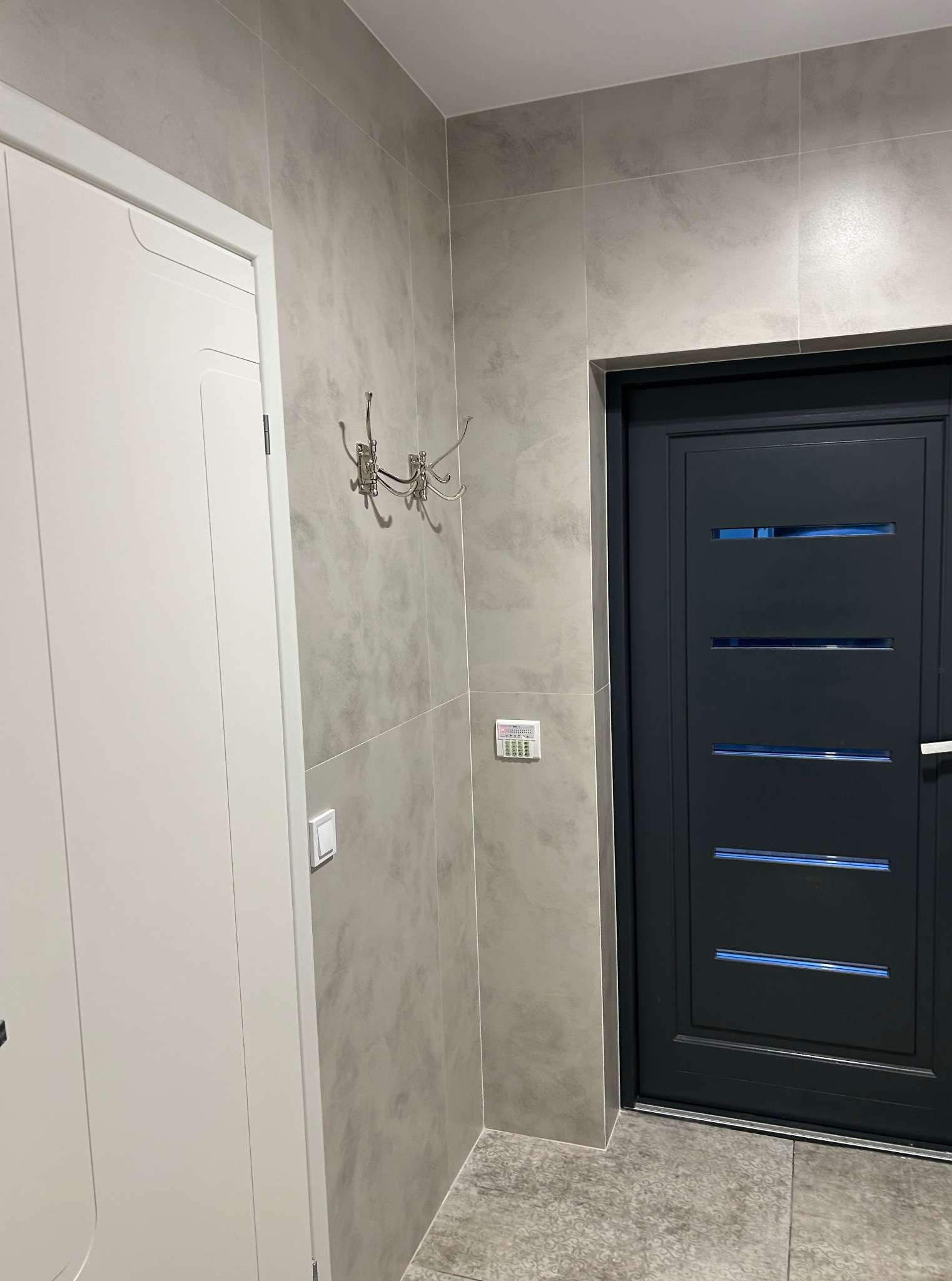 Nowoczesny przedpokój z szarą, teksturowaną płytką na ścianach, białą szafą i ciemnoszarymi drzwiami z poziomymi przeszkleniami. W tle widoczny panel sterowania.