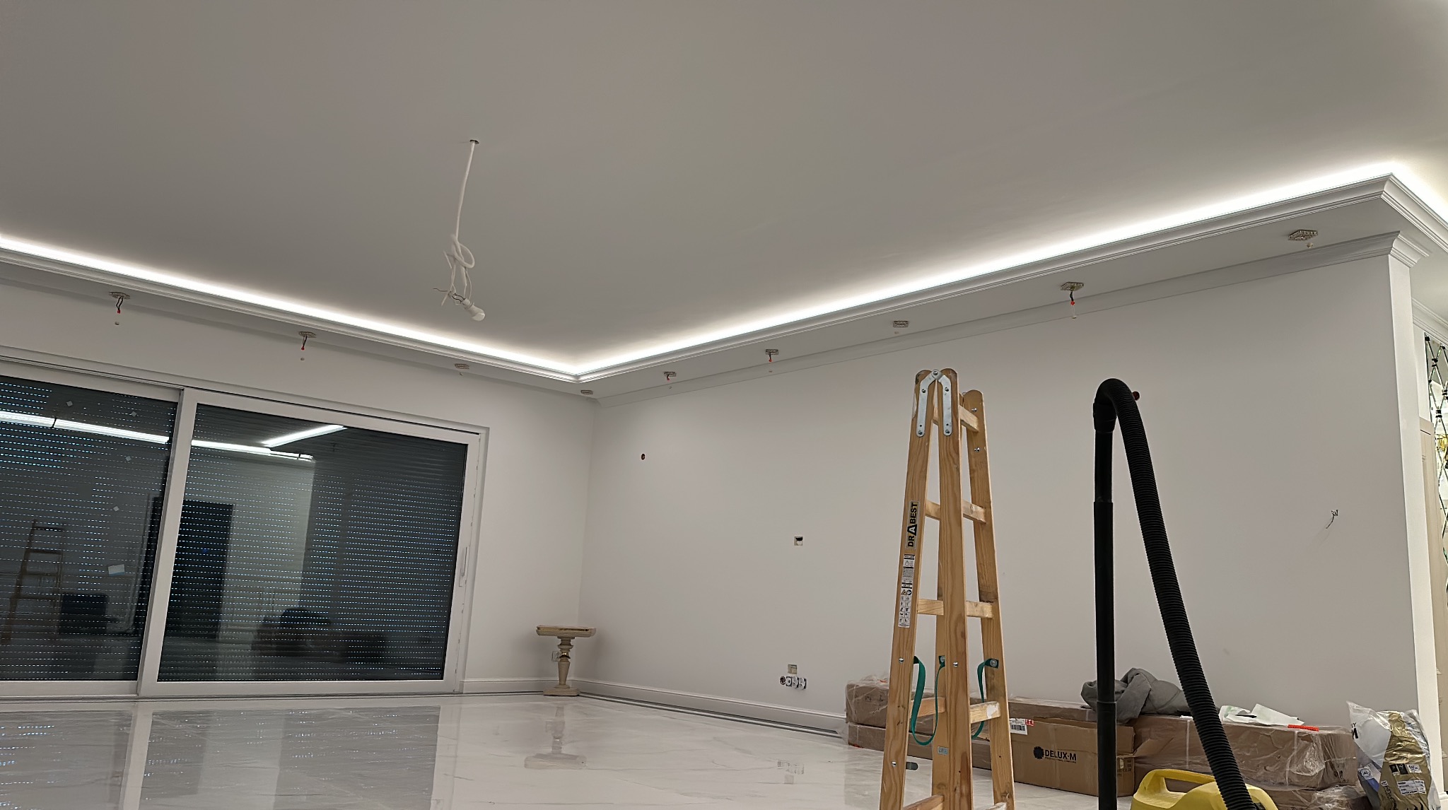 Jasne wnętrze w trakcie wykańczania z oświetleniem LED podsufitowym, drewnianą drabiną i odkurzaczem. Marmurowa podłoga odbija światło, tworząc elegancki efekt.