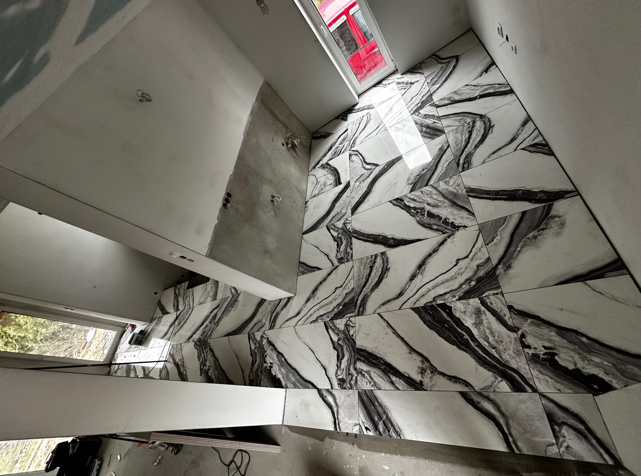 Ukończona podłoga z płytek imitujących marmur w łazience z oknem dachowym. Częściowo wykończone ściany. Widoczna instalacja elektryczna.