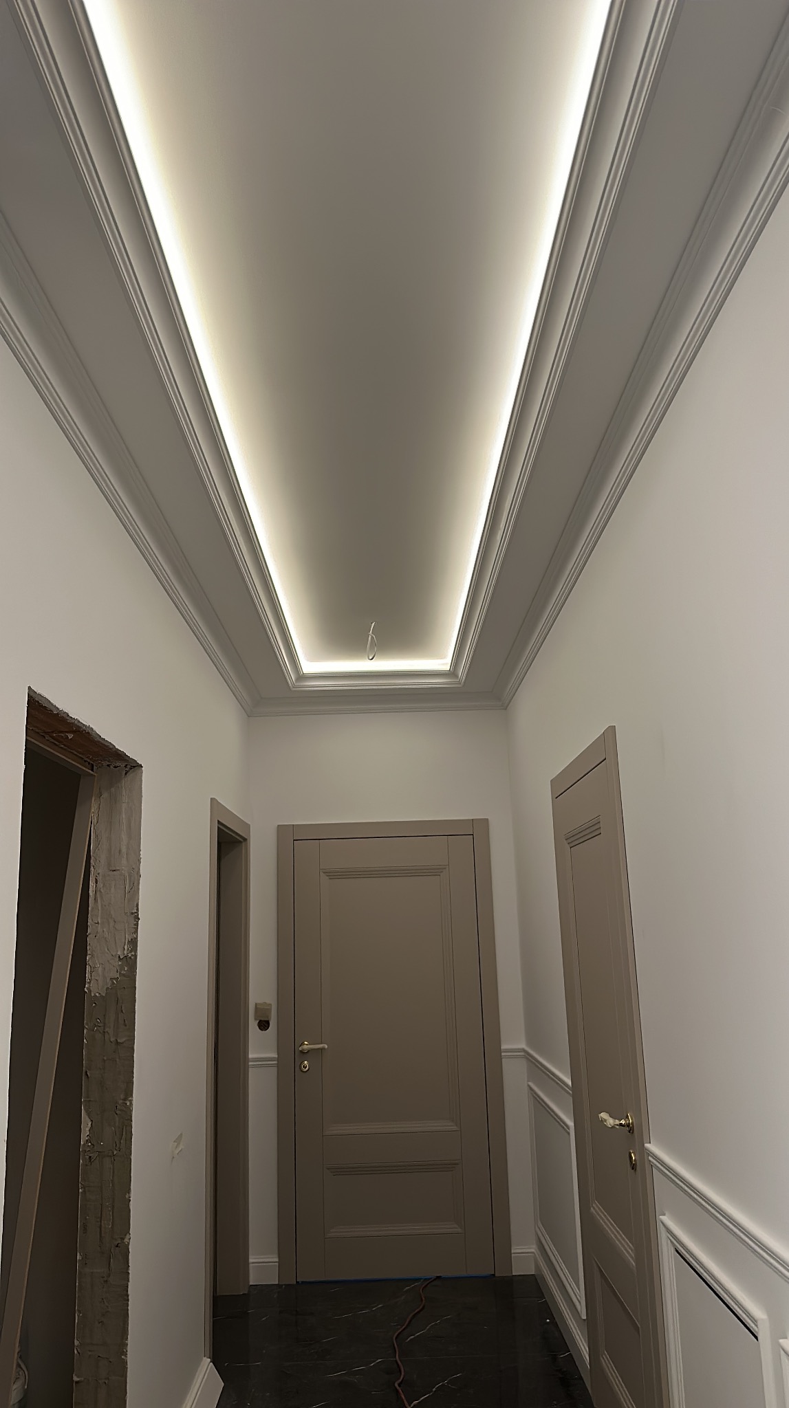 Korytarz po remoncie: białe ściany z dekoracyjną sztukaterią, podświetlany sufit kasetonowy, dwie szare drzwi, czarna marmurowa podłoga, w tle widoczny fragment ściany w trakcie prac.