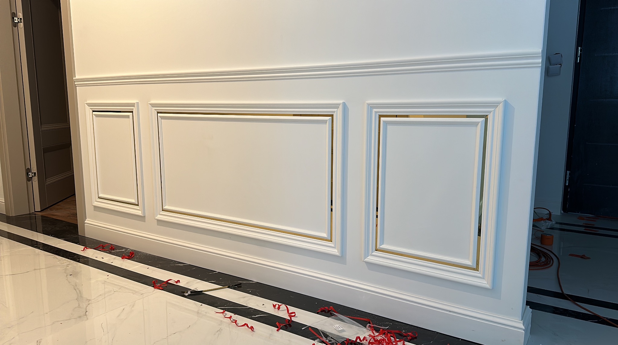 Eleganckie wykończenie ściany z białymi panelami i złotymi akcentami, widoczne detale listew przypodłogowych i marmurowej podłogi w paski.