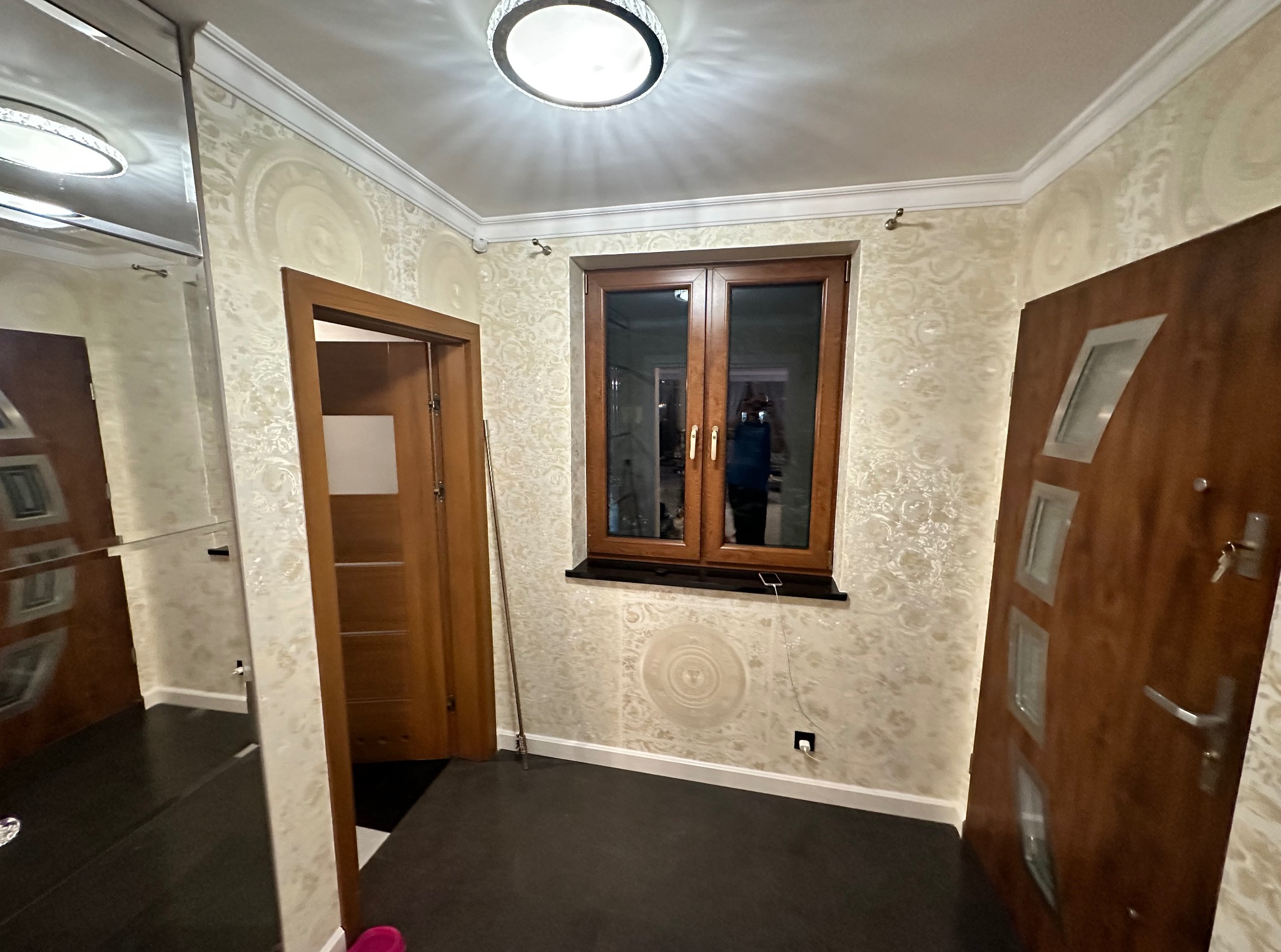 Eleganckie wykończenie wnętrza: wzorzyste tapety w kremowych odcieniach, drewniane okno i drzwi, nowoczesna lampa sufitowa, ciemna podłoga. Detal: listwa przypodłogowa.