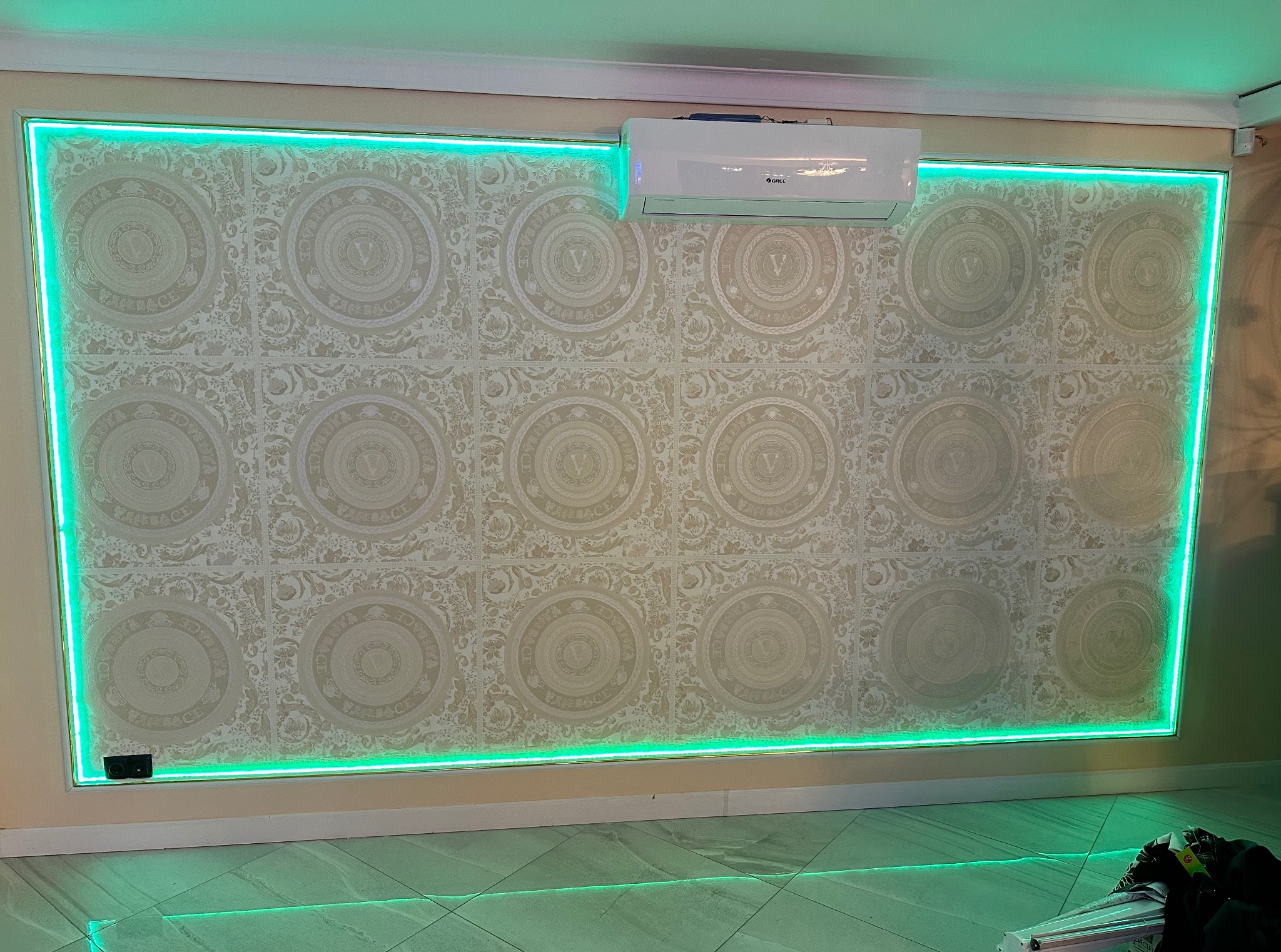 Ściana z dekoracyjną tapetą w kremowych odcieniach, obramowana zielonym oświetleniem LED. Na górze widoczny klimatyzator. Nietypowe wykończenie wnętrza.