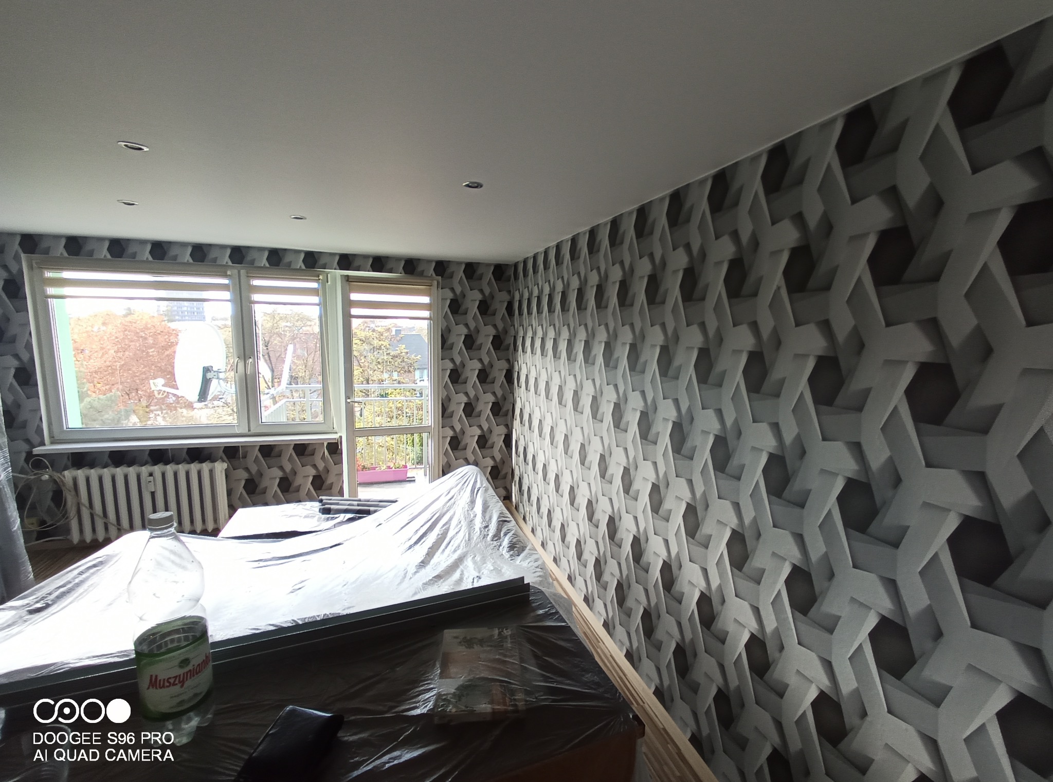 Pokój w trakcie remontu z geometryczną tapetą na ścianie. Podłoga i meble zabezpieczone folią malarską. Widoczne okno i grzejnik. W tle puszka po konserwie.