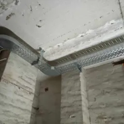 Metalowe korytko kablowe zamontowane pod nierównym, betonowym sufitem w surowym wnętrzu. Widoczne przewody elektryczne i fragmenty ścian z ubytkami tynku.