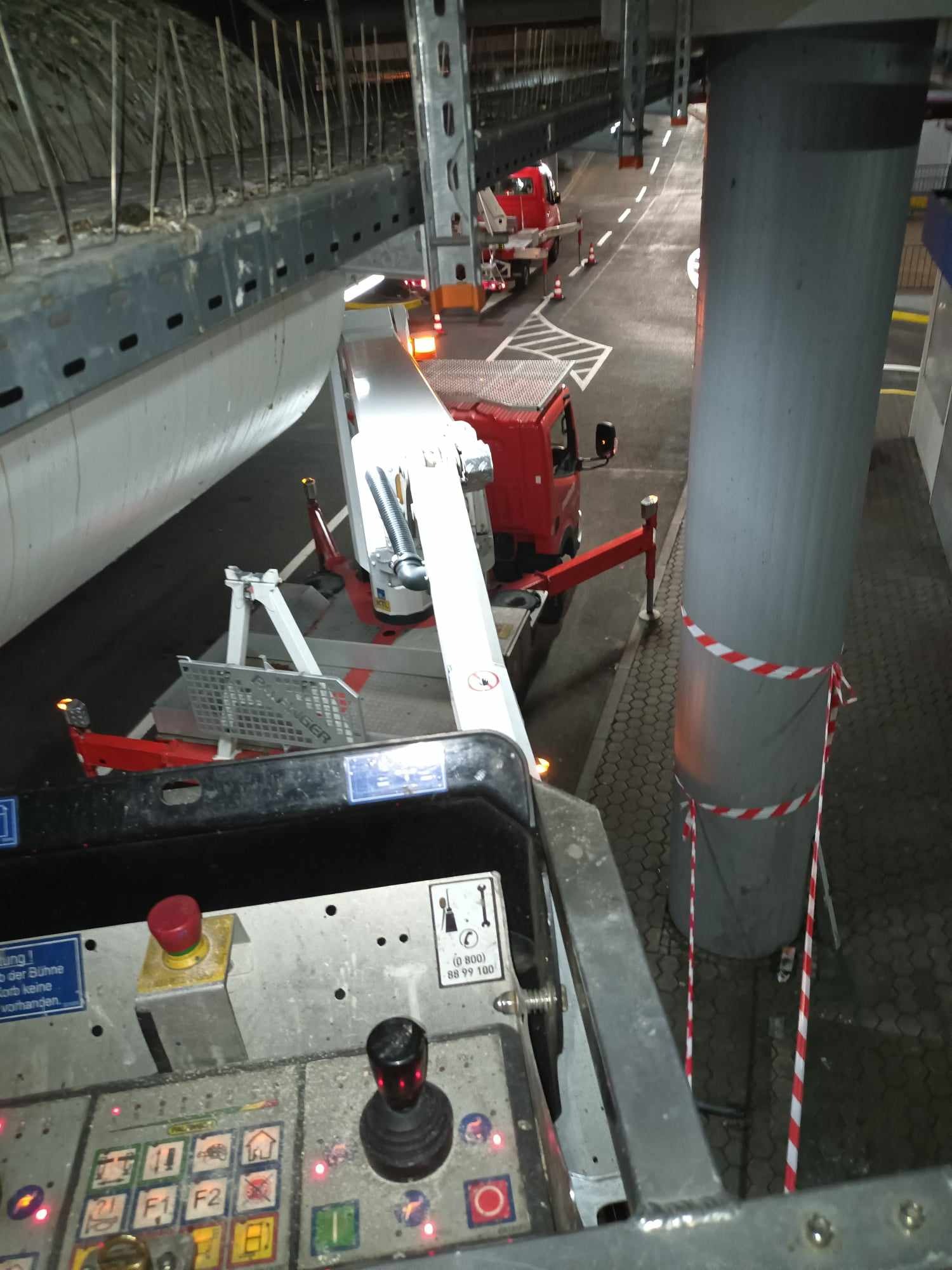 Widok z góry na platformę roboczą podnośnika koszowego, z widocznym panelem sterowania, betonową konstrukcją stropu z wystającymi prętami zbrojeniowymi, oraz dwoma czerwonymi samochodami na parkingu.