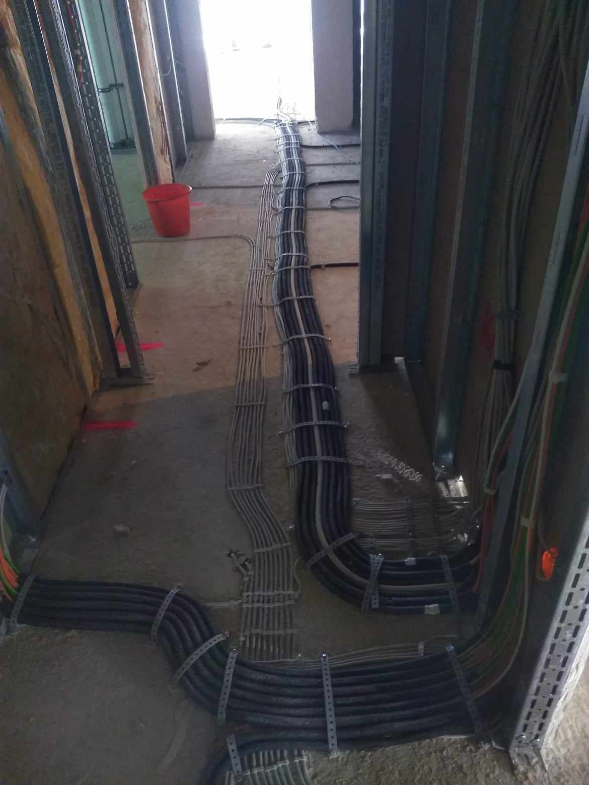 Wiązki przewodów elektrycznych ułożone w korycie na podłodze w surowym stanie, zabezpieczone metalowymi uchwytami, widoczne elementy konstrukcji ścian z profili stalowych.