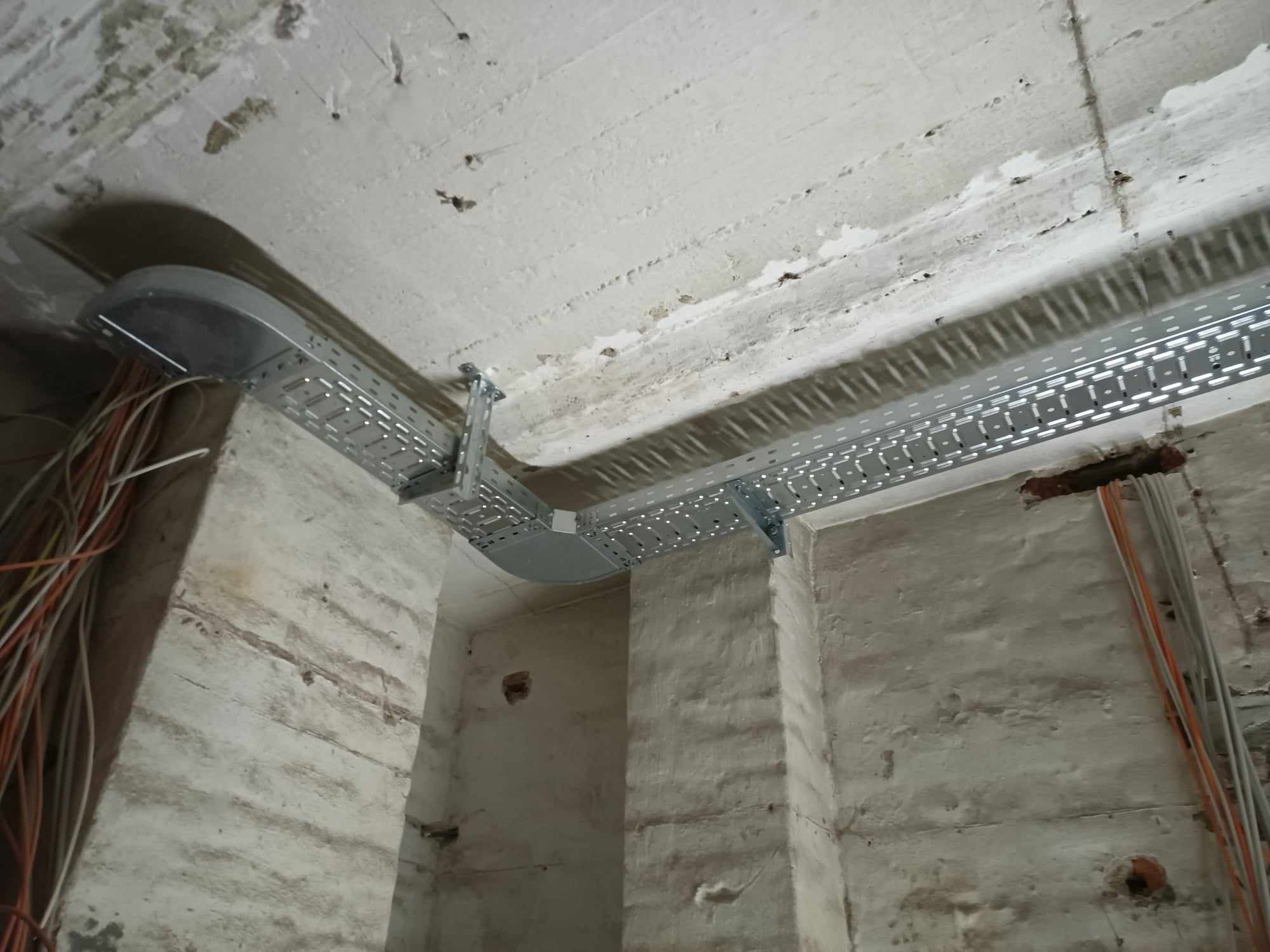 Metalowe korytko kablowe zamontowane pod nierównym, betonowym sufitem w surowym wnętrzu. Widoczne przewody elektryczne i fragmenty ścian z ubytkami tynku.