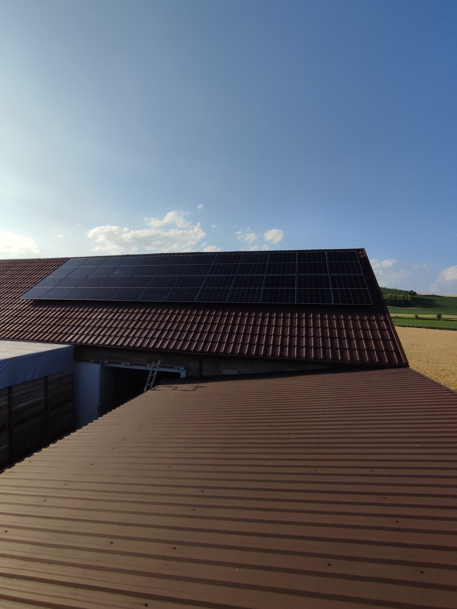 Panele słoneczne na brązowym dachu z dachówki ceramicznej, widok z innego dachu z blachy trapezowej, błękitne niebo w tle.