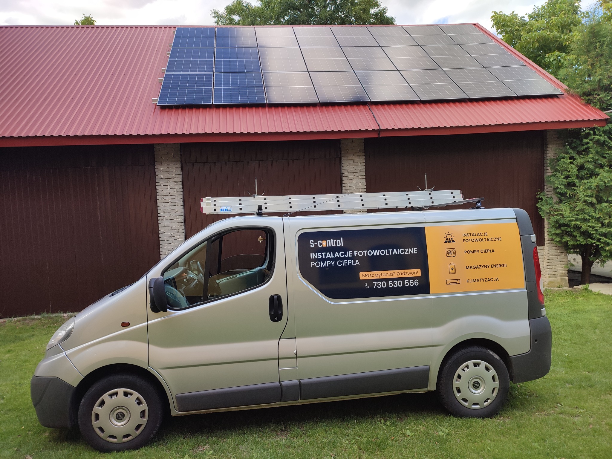 Panel słoneczny na czerwonym dachu garażu, srebrny samochód dostawczy z reklamą firmy instalującej panele fotowoltaiczne i pompy ciepła na trawniku.