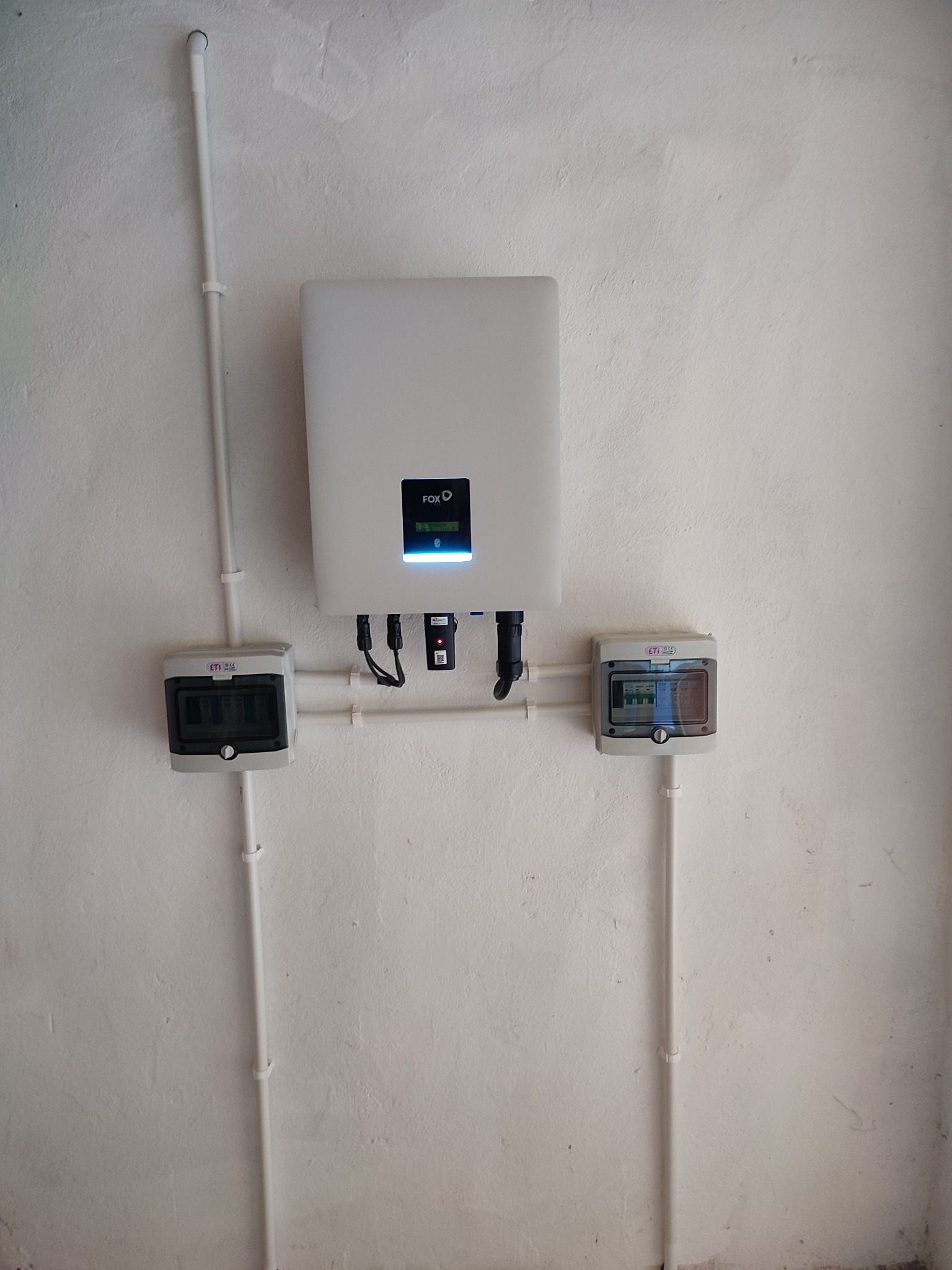 Biały inwerter z podłączonymi przewodami i dwoma skrzynkami bezpiecznikowymi na białej, lekko chropowatej ścianie wewnątrz budynku.