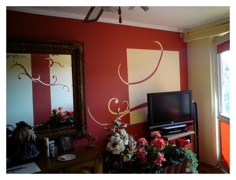 Pokój po malowaniu ścian z dekoracyjnym motywem w kolorach czerwieni i beżu, widoczny telewizor i lustro w ozdobnej ramie.