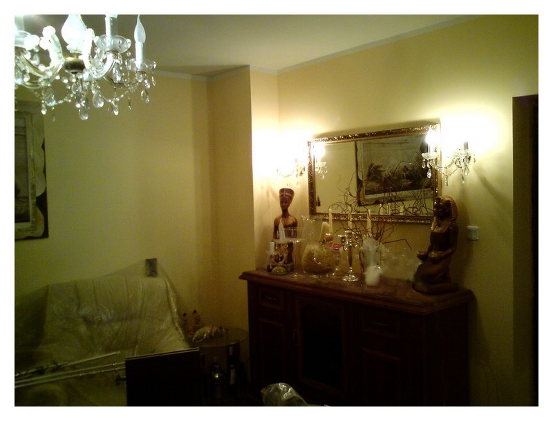 Salon w ciepłej tonacji z kryształowym żyrandolem, komodą z dekoracjami w stylu egipskim i lustrem w złotej ramie, sofa przykryta folią ochronną.