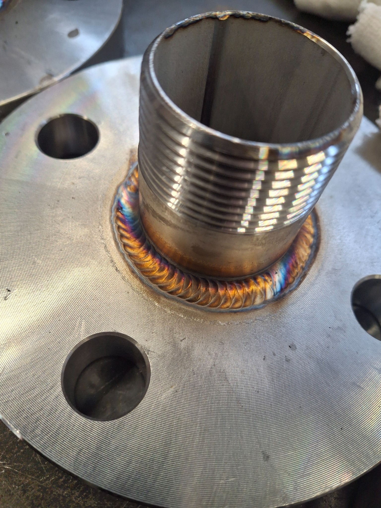 Metalowa flansza z przyspawaną rurą gwintowaną, widoczne tęczowe zabarwienie spoiny, świadczące o jakości wykonania.