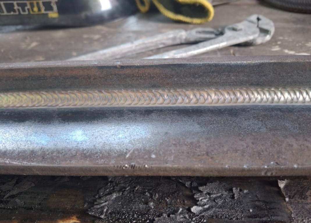 Precyzyjne spoiwo na metalowym profilu kątowym, wykonane metodą spawania MIG/MAG. Widoczne narzędzia w tle i ślady obróbki na powierzchni.