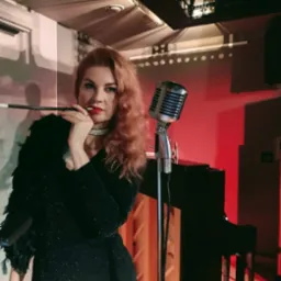 Piosenkarka w czarnej sukni z boa z piór, z ustnikiem w ustach, stoi obok mikrofonu retro i pianina, w tle czerwone światło i dekoracja imitująca miasto.