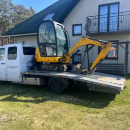 Żółta minikoparka JCB załadowana na białą lawetę przed domem z balkonem i zielonym dachem, w tle las.