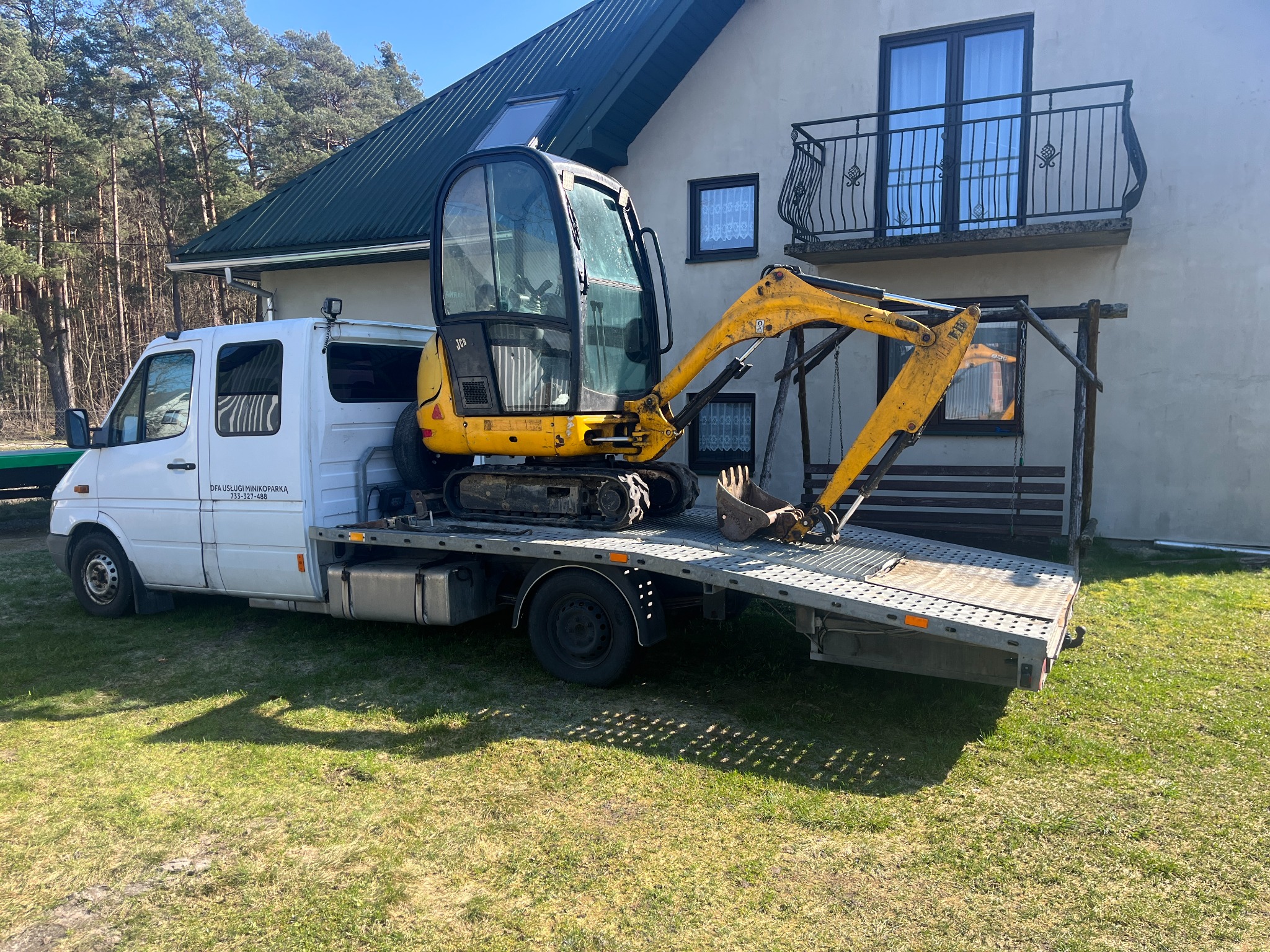 Żółta minikoparka JCB załadowana na białą lawetę przed domem z balkonem i zielonym dachem, w tle las.