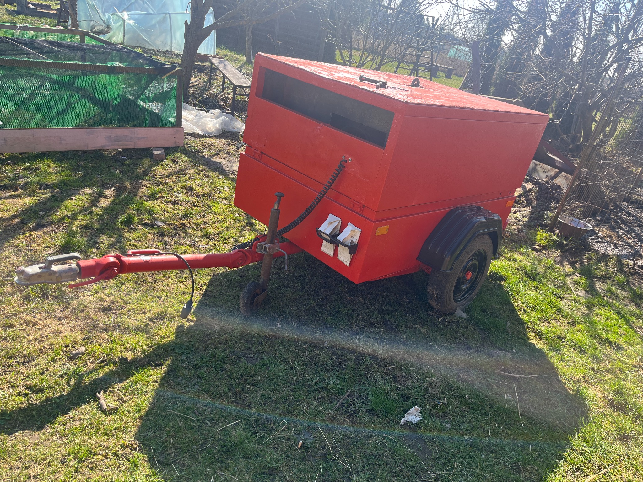 Czerwona, skrzyniowa przyczepa na trawie, z widocznym zaczepem i kablem zasilającym, sugerująca transport sprzętu telekomunikacyjnego lub energetycznego, w tle szklarnia i drzewa bez liści.