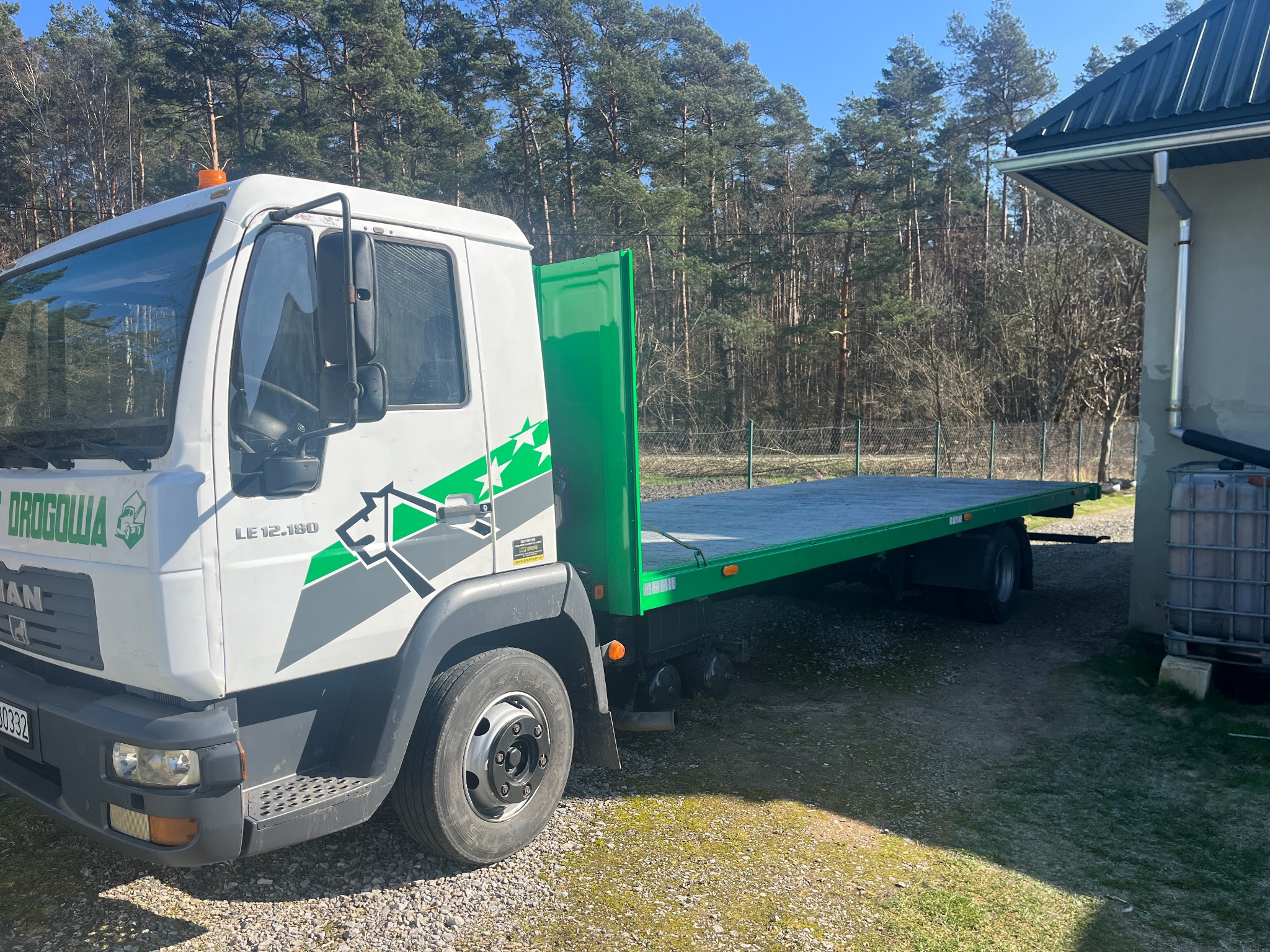 Biała ciężarówka MAN z zieloną platformą transportową, ozdobiona logo firmy z motywem konia, zaparkowana na żwirowym podłożu, na tle budynku i wysokich drzew.