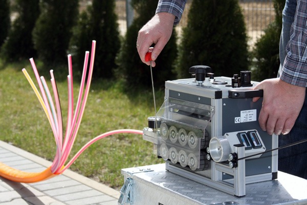 Pracownik używa specjalistycznej maszyny do obróbki kabli światłowodowych na zewnątrz, widoczne kolorowe włókna światłowodowe wystające z pomarańczowej osłony.