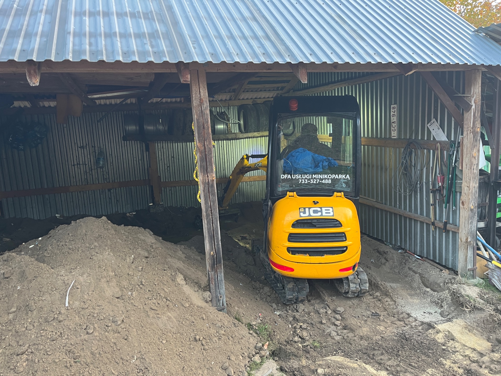Żółta mini koparka JCB w zadaszonej przestrzeni roboczej, obok pryzma ziemi, widoczny numer telefonu firmy na szybie kabiny.