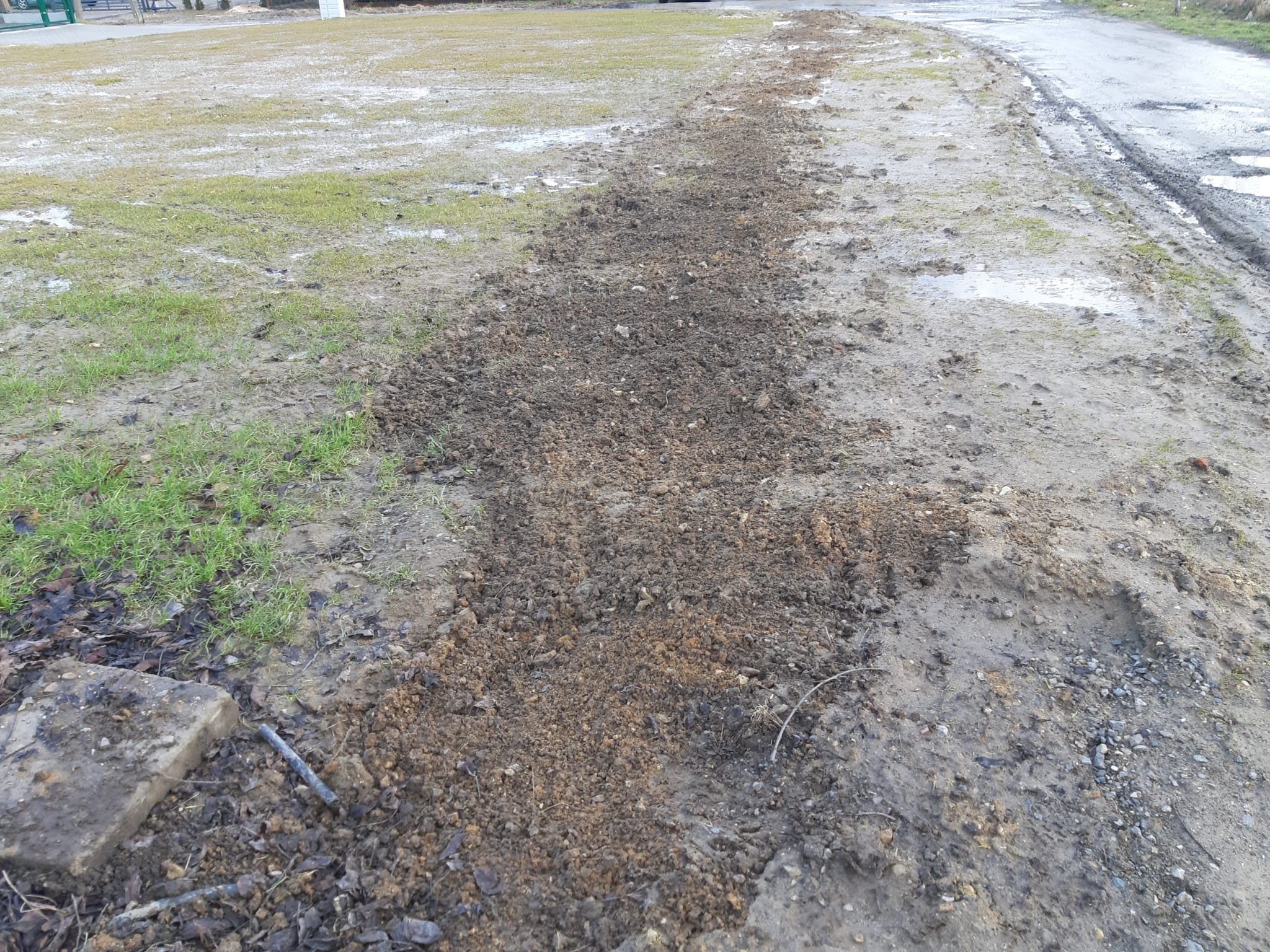 Świeżo wykopany, wąski rów w ziemi na podmokłym terenie, widoczne ślady błota i trawy, obok fragment betonowej płyty i wystające metalowe rurki.