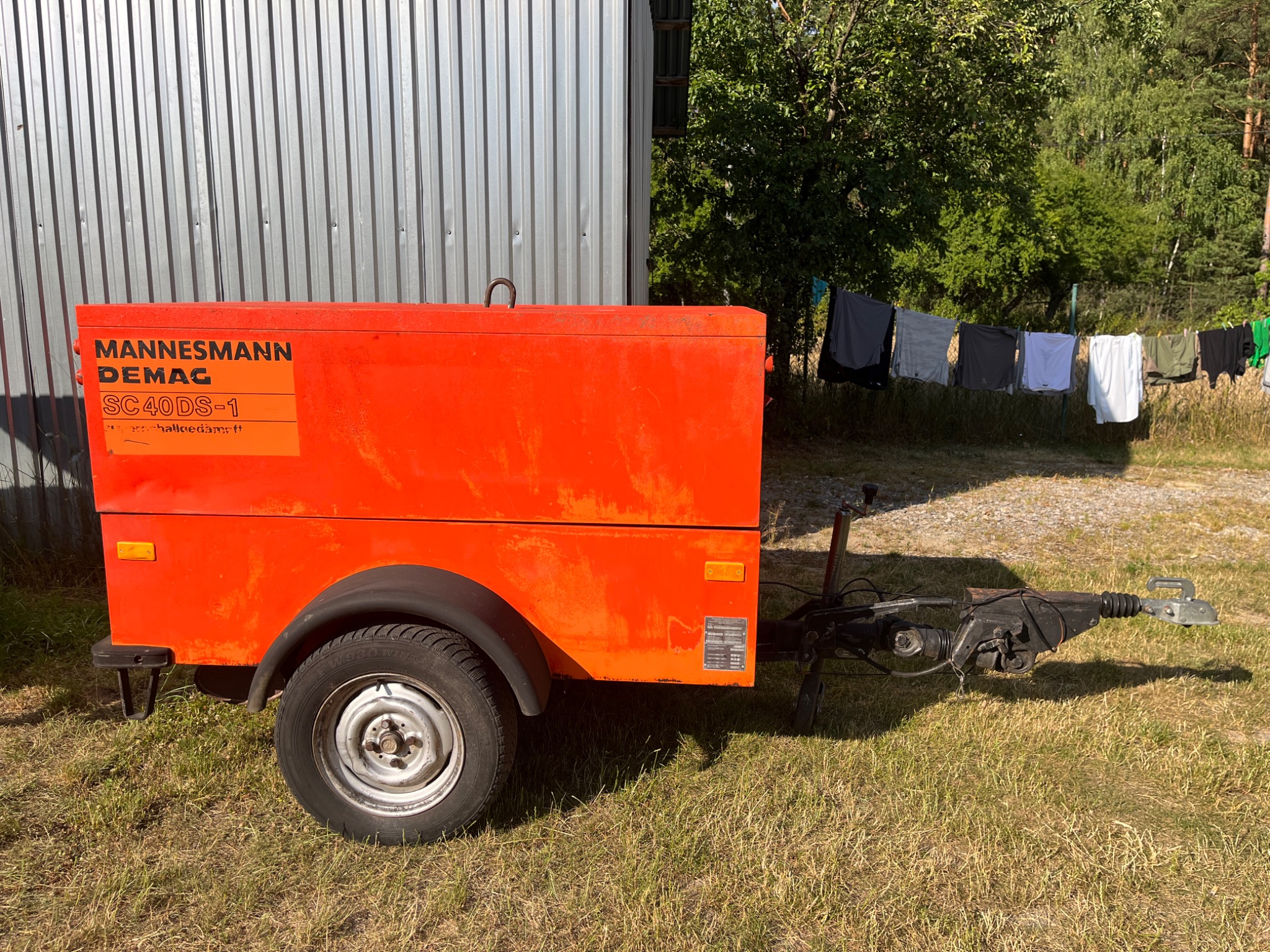 Pomarańczowa przyczepa kompresora Mannesmann Demag SC40DS-1 na trawie, z blachy falistej w tle i praniem suszącym się na sznurku w oddali.