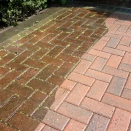 Częściowo umyta kostka brukowa ułożona w jodełkę, z widocznymi chwastami. Po lewej stronie kostka jest mokra i brudna, po prawej sucha i czysta.