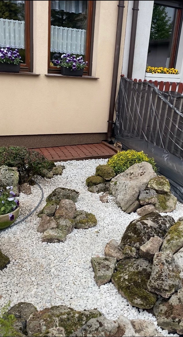 Ogród z białym żwirem, kamieniami porośniętymi mchem i roślinami. Na parapecie okiennym kwiaty w donicach. Folia ochronna na płocie w tle.