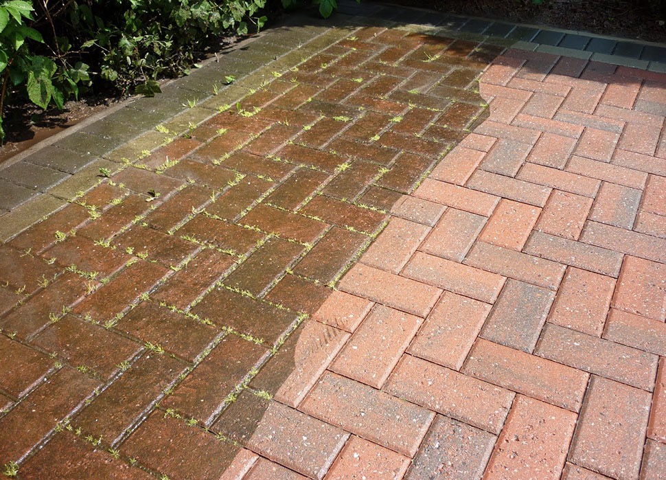 Częściowo umyta kostka brukowa ułożona w jodełkę, z widocznymi chwastami. Po lewej stronie kostka jest mokra i brudna, po prawej sucha i czysta.