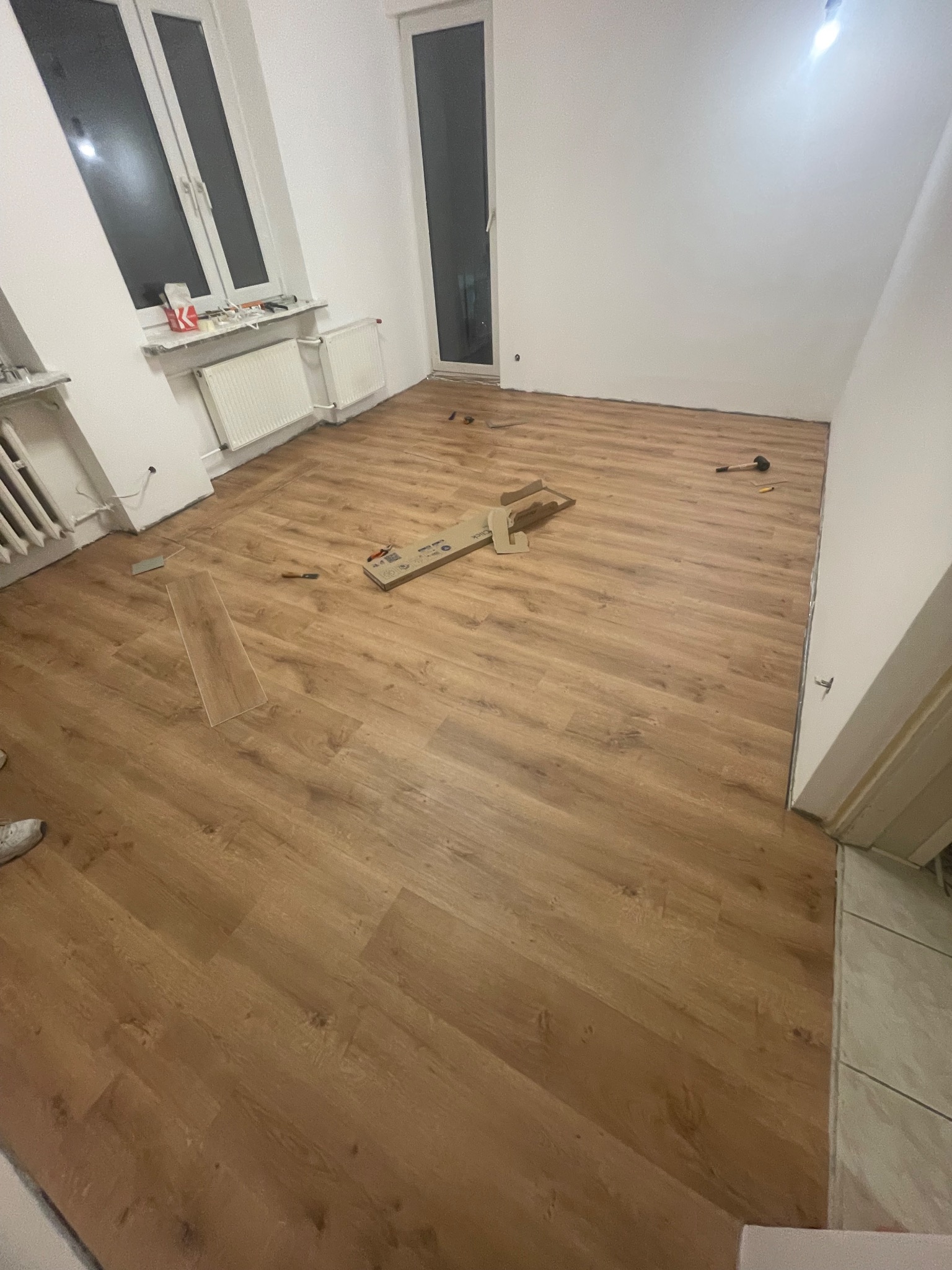 Układanie paneli podłogowych w jasnym pokoju. Widoczne narzędzia i fragment kartonu po panelach. Częściowo ułożona podłoga z paneli imitujących drewno.