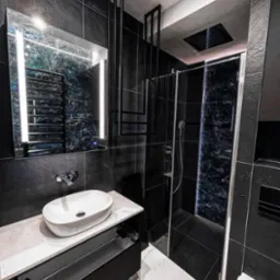 Nowoczesna łazienka w ciemnej tonacji z czarnymi płytkami imitującymi kamień, owalną białą umywalką nablatową, podświetlanym lustrem i kabiną prysznicową z dekoracyjnym panelem. Dodatkowy element...