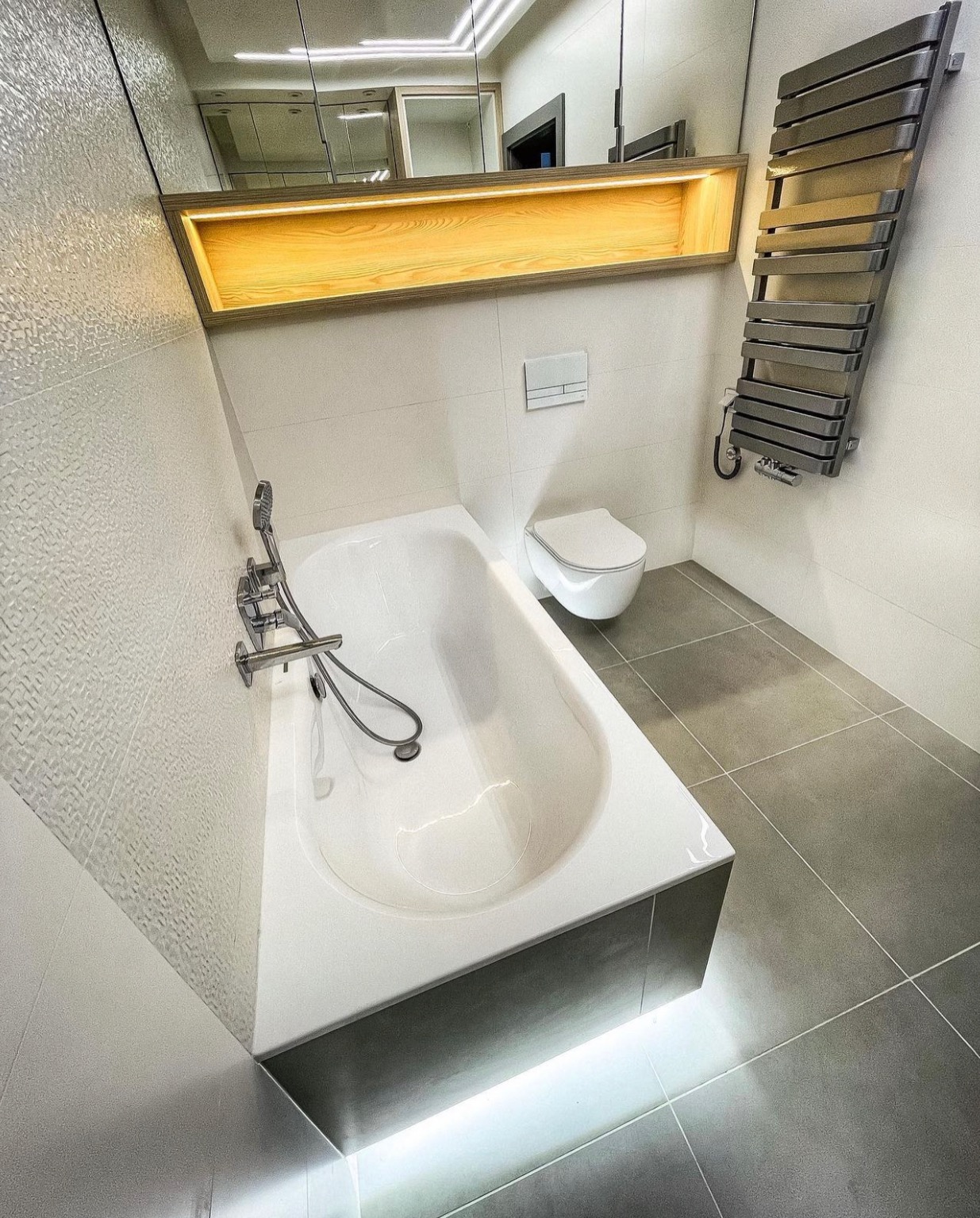 Nowoczesna łazienka z białą wanną podświetloną od dołu, szara podłoga z płytek, białe płytki na ścianach, grzejnik drabinkowy w kolorze grafitowym oraz podświetlana drewniana półka nad toaletą.