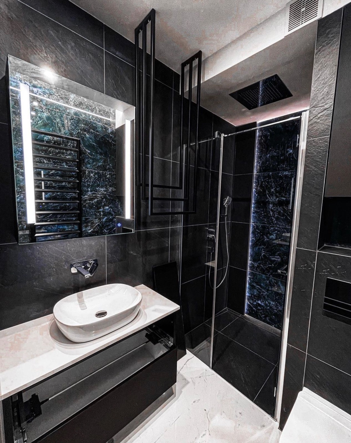 Nowoczesna łazienka w ciemnej tonacji z czarnymi płytkami imitującymi kamień, owalną białą umywalką nablatową, podświetlanym lustrem i kabiną prysznicową z dekoracyjnym panelem. Dodatkowy element...