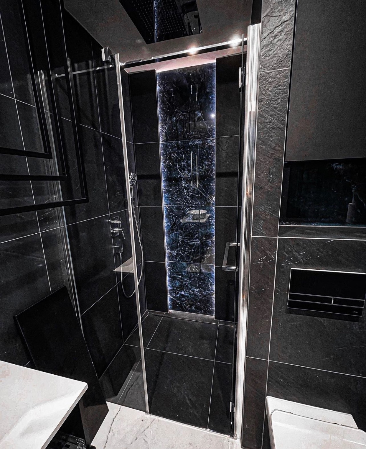 Nowoczesna kabina prysznicowa z czarnymi płytkami imitującymi kamień i panelem dekoracyjnym z efektem marmuru podświetlonym od wewnątrz, chromowane wykończenia, biała umywalka w rogu.
