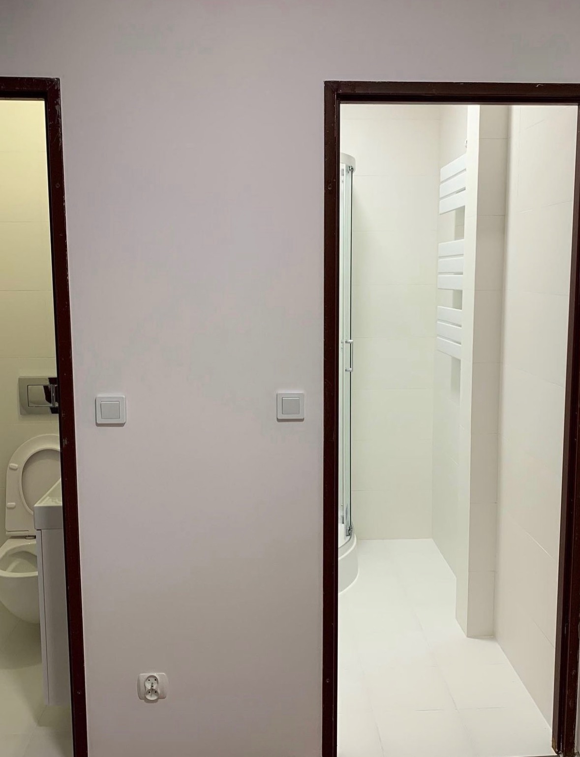 Widok na dwie łazienki przez otwarte drzwi: po lewej toaleta i umywalka, po prawej kabina prysznicowa i grzejnik drabinkowy, białe płytki i ściany, brązowe ramy drzwi.