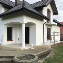 Nowo wykończona biała elewacja domu z ciemną dach&oacute;wką, widoczne ślady po malowaniu na tarasie i narzędzia malarskie, sugerujące świeżo zakończone prace.