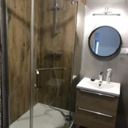 Nowoczesna łazienka z kabiną prysznicową narożną, umywalką z szafką w kolorze drewna i okrągłym lustrem podświetlanym lampą.