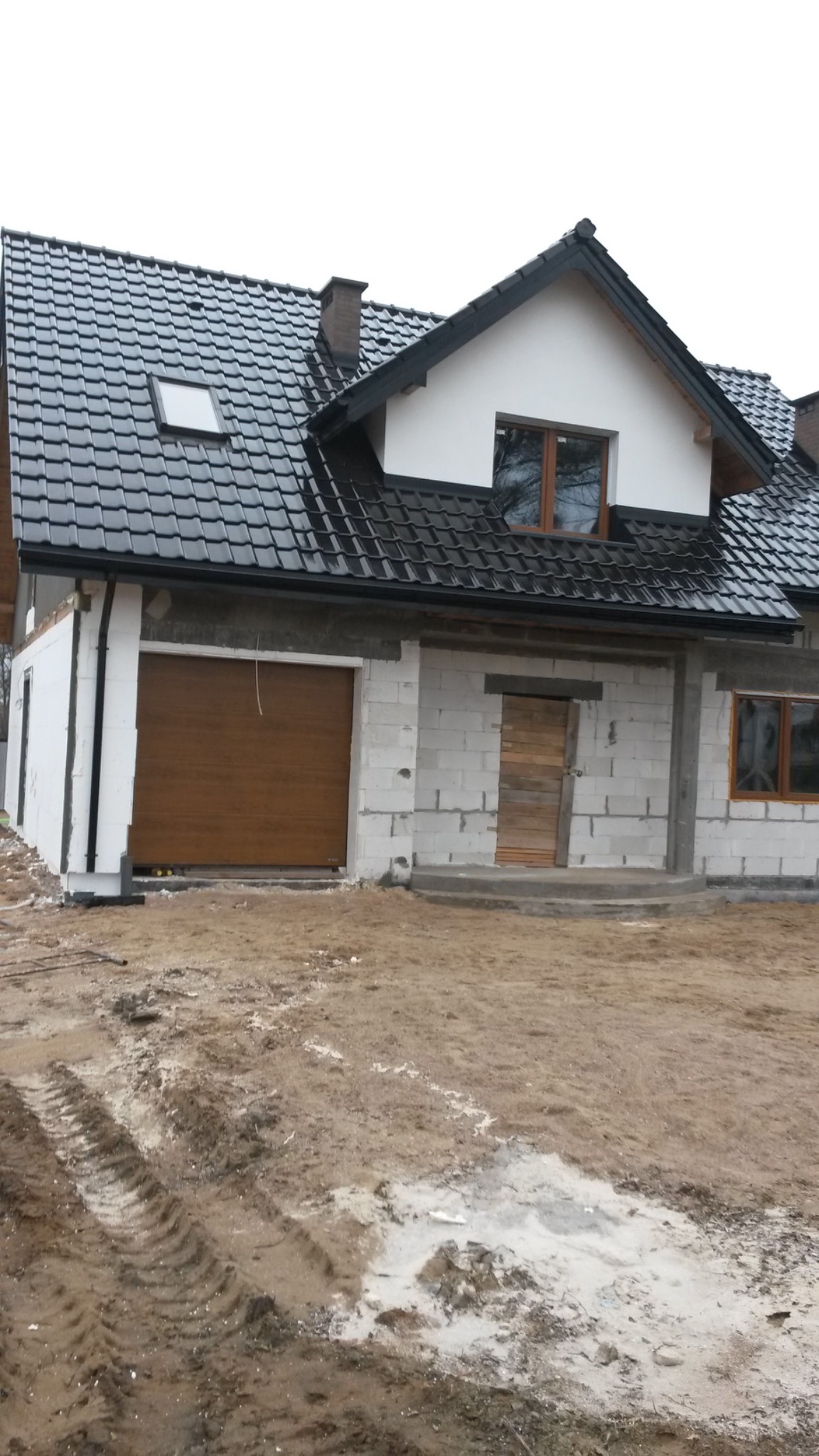 Dom w trakcie budowy: widoczna czarna dachówka, brama garażowa z brązowego drewna i ściany z białego pustaka.