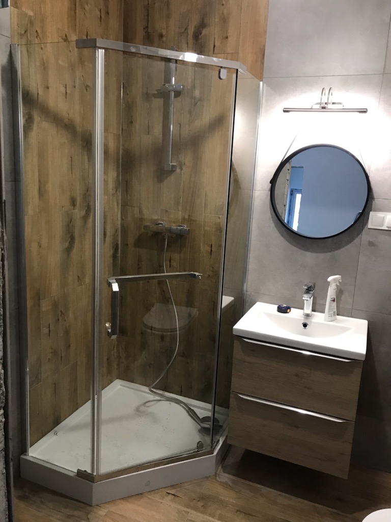 Nowoczesna łazienka z kabiną prysznicową narożną, umywalką z szafką w kolorze drewna i okrągłym lustrem podświetlanym lampą.