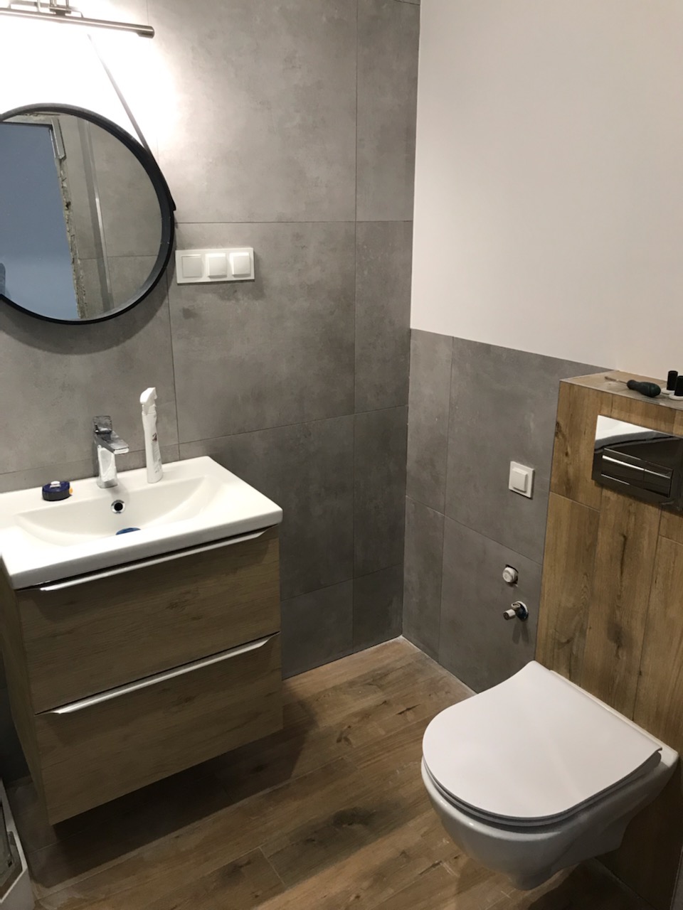 Nowoczesna łazienka z szarą glazurą, drewnopodobną podłogą i wiszącą toaletą, widoczne elementy instalacji wodnej oraz okrągłe lustro z czarną ramą.