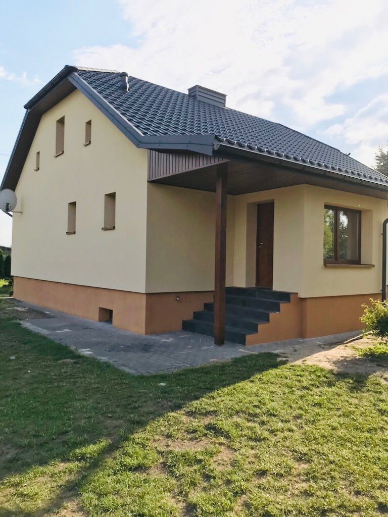 Dom jednorodzinny po remoncie elewacji, z nowym dachem z blachodachówki, ocieplony, pomalowany na beżowo z brązowym cokołem, widoczne schody wejściowe z ciemnych płytek.