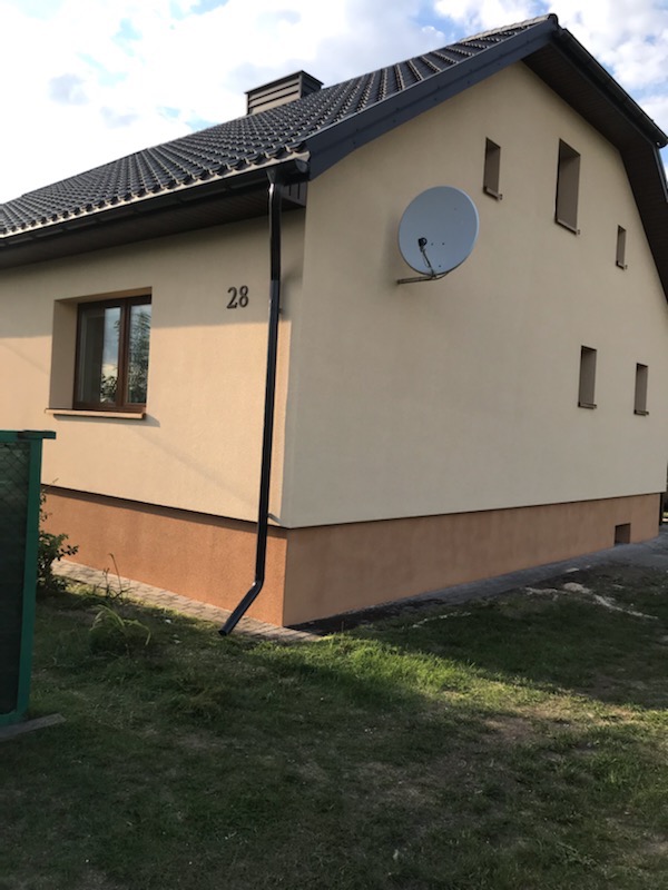 Dwukondygnacyjny dom z jasnobeżową elewacją i ciemnobrązowym pasem u dołu, widoczny numer domu '28' oraz antena satelitarna na ścianie, dach pokryty ciemną dachówką