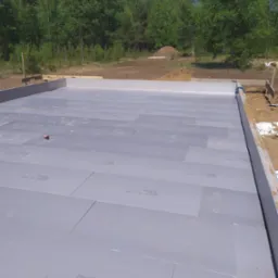 Przygotowany fundament pod budynek, pokryty szarymi płytami izolacyjnymi Synthos XPS Prime S, widoczne drewniane szalunki i teren budowy z drzewami w tle.